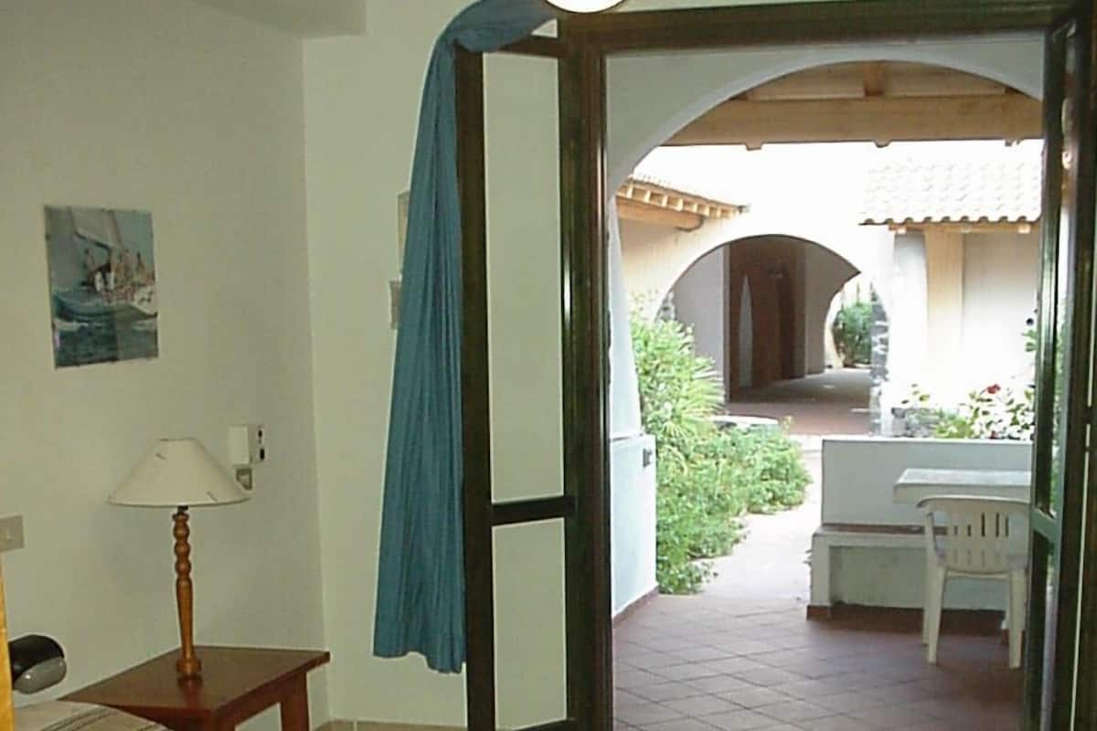 A bedroom with a bed, a nightstand, a lamp, and a view of an outdoor patio with a table and chair.
