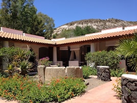 Um pátio com telhado de azulejos, pilares de pedra e diversas plantas, incluindo cravos-de-defunto e agave.