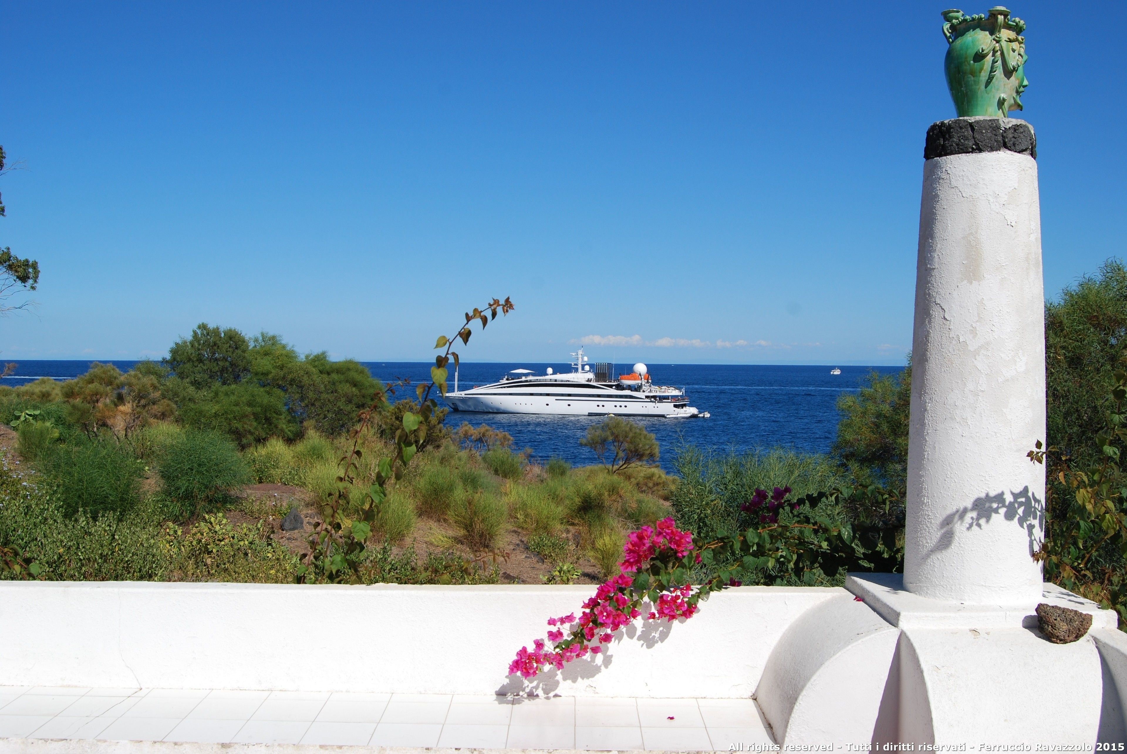 A white column with a green top, a white wall with a pink flower arrangement, and a yacht in the background.