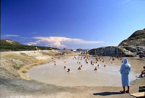 Un spa geotérmico con una piscina grande rodeada de terreno rocoso y un cielo azul despejado.