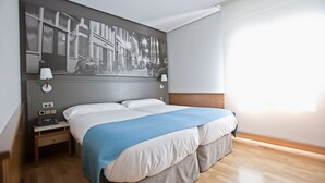 A modern hotel room with a large bed, two bedside tables, a wall-mounted lamp, and a large black-and-white cityscape painting. - Hotel Don Carlos (Huarte)