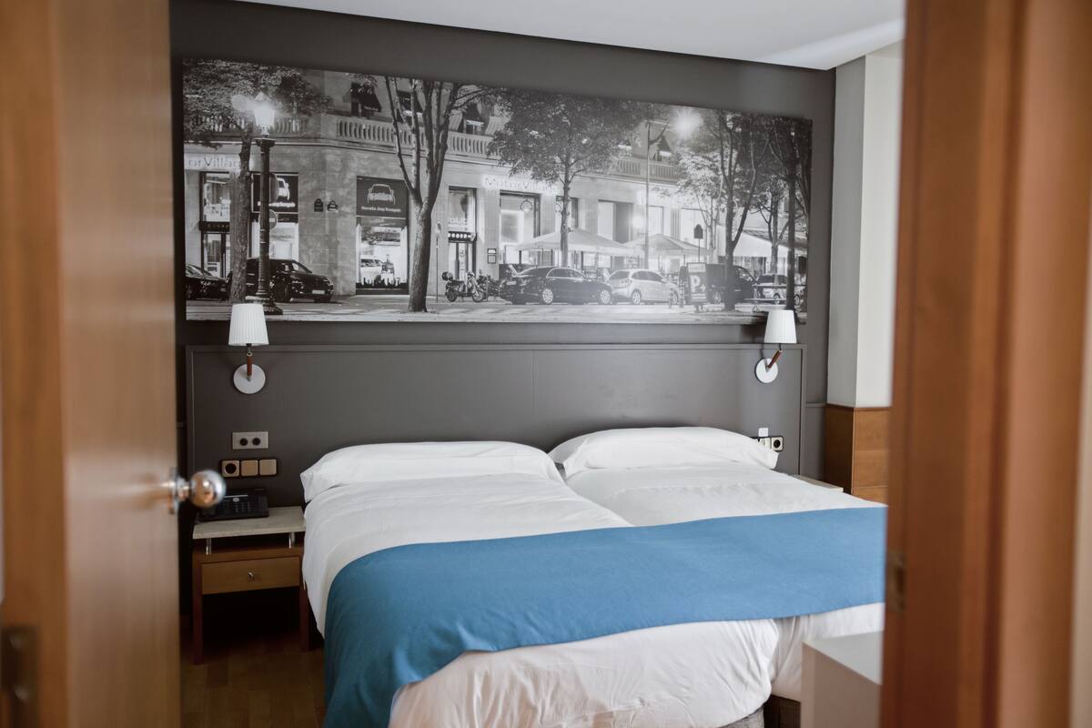 A hotel room with a bed, a nightstand, a lamp, and a wall-mounted painting of a street scene.