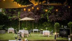 Un área de comedor al aire libre con mesas y sillas bajo un toldo, rodeada de exuberante follaje y un cartel que dice 'LA ESTIVA RESTAURANTE'.