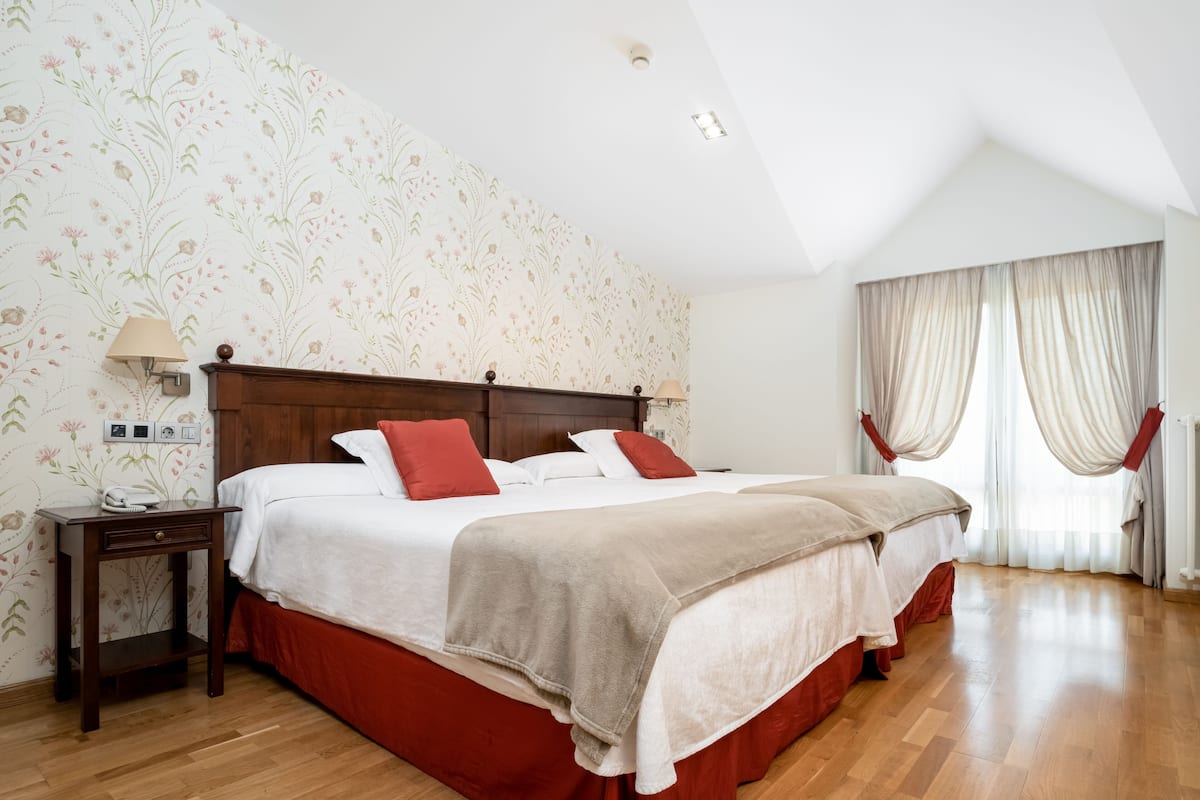 Ein Schlafzimmer mit einem hölzernen Bett, rot-weiß gestreifter Bettwäsche, blumigem Tapetenmuster, einem Nachttisch und einem Fenster mit Vorhängen.