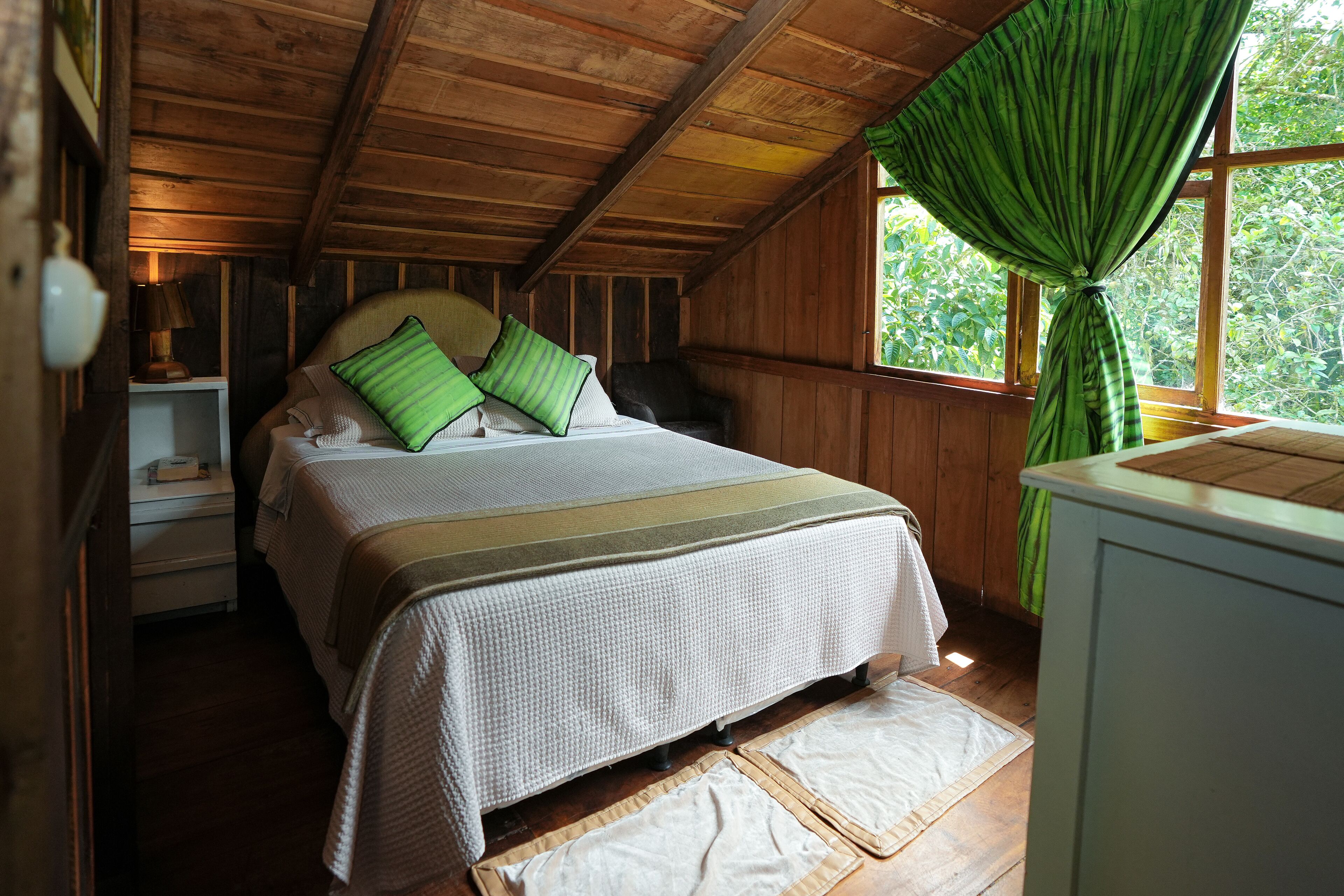 Un dormitorio de cabaña de madera con una cama, almohadas, una mesita de noche y una ventana con cortinas.
