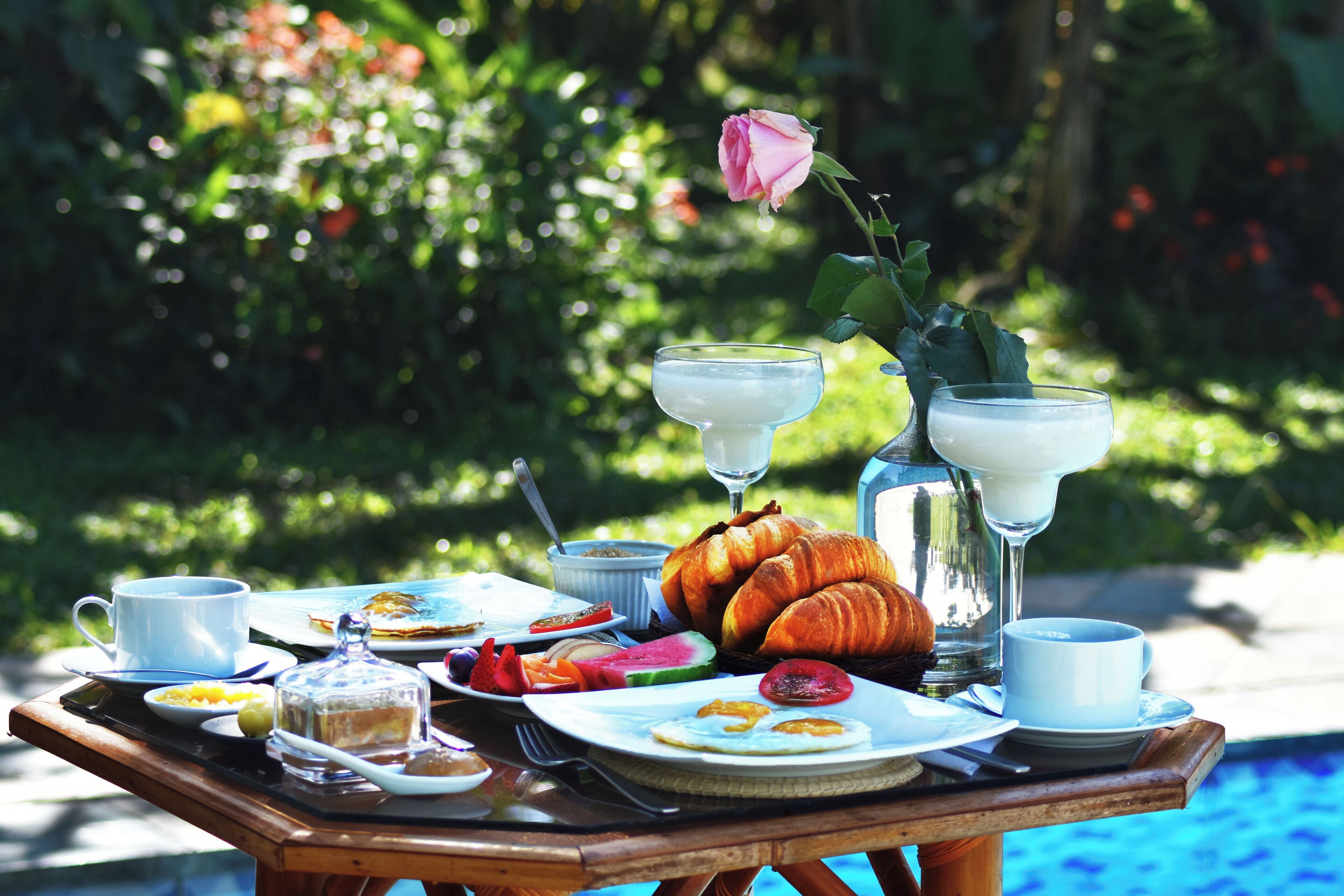 A breakfast table with croissants, eggs, and fruit, set by a poolside.