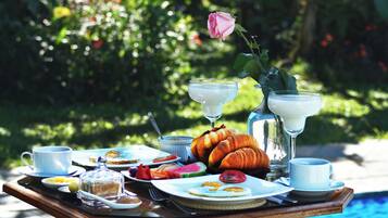 A breakfast table with croissants, eggs, and fruit, set by a poolside.