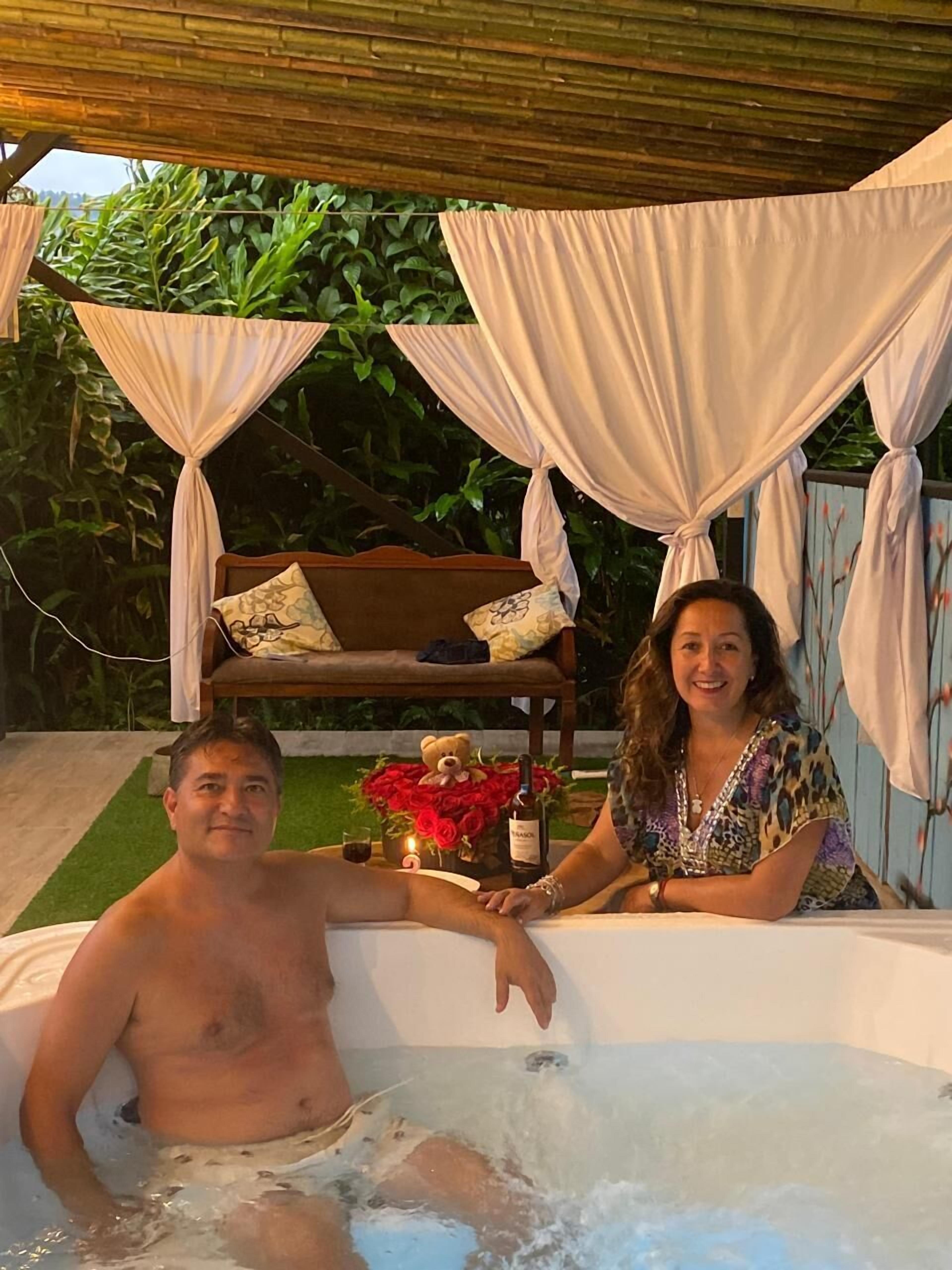 Una pareja en un jacuzzi con un banco de madera y cojines al fondo, rodeada de follaje.