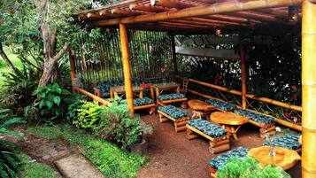 An outdoor seating area with bamboo furniture and a pergola structure, surrounded by lush greenery.