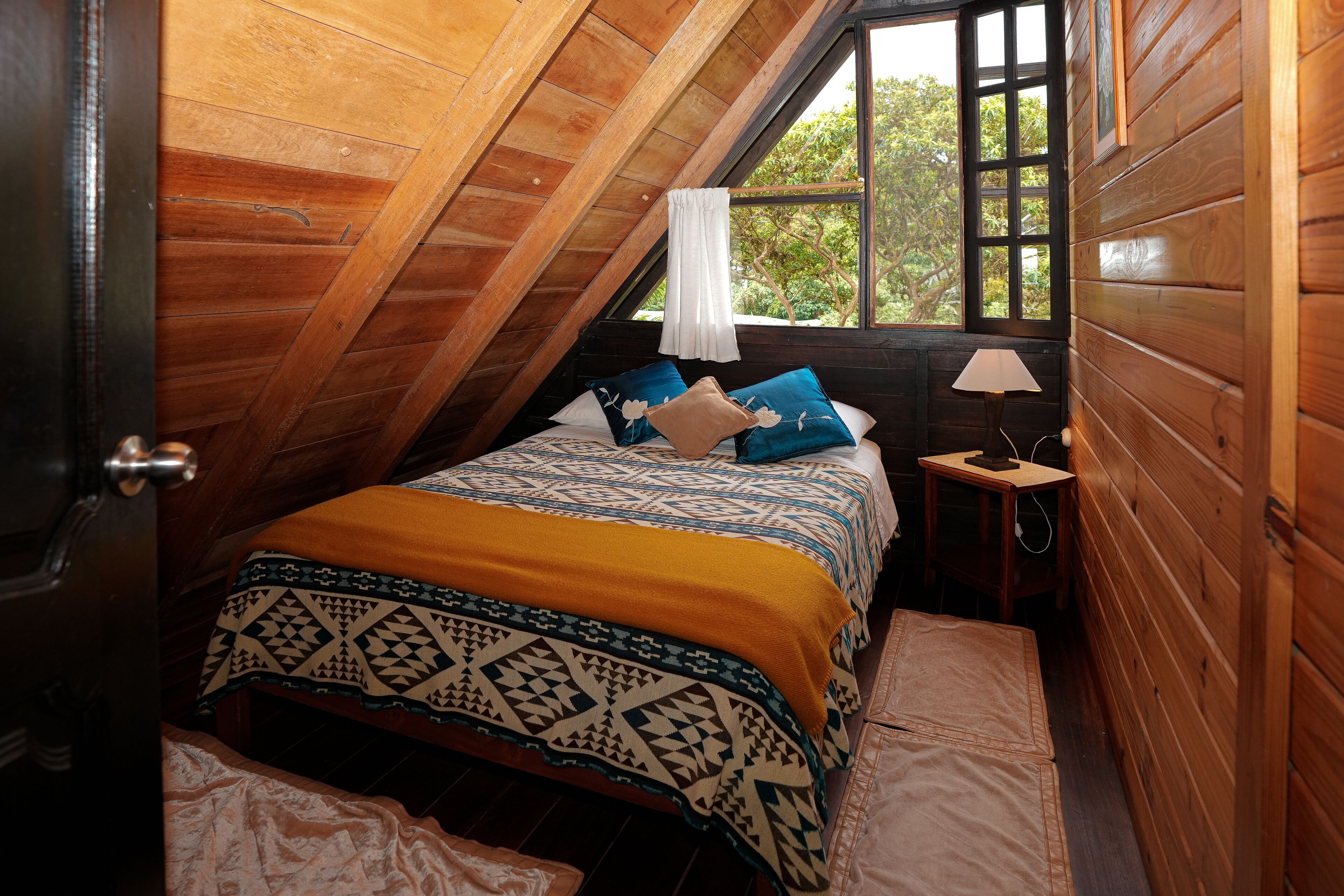 A cozy attic bedroom with a wooden sloped ceiling, a bed with patterned bedding, a nightstand with a lamp, and a window with a view of trees.