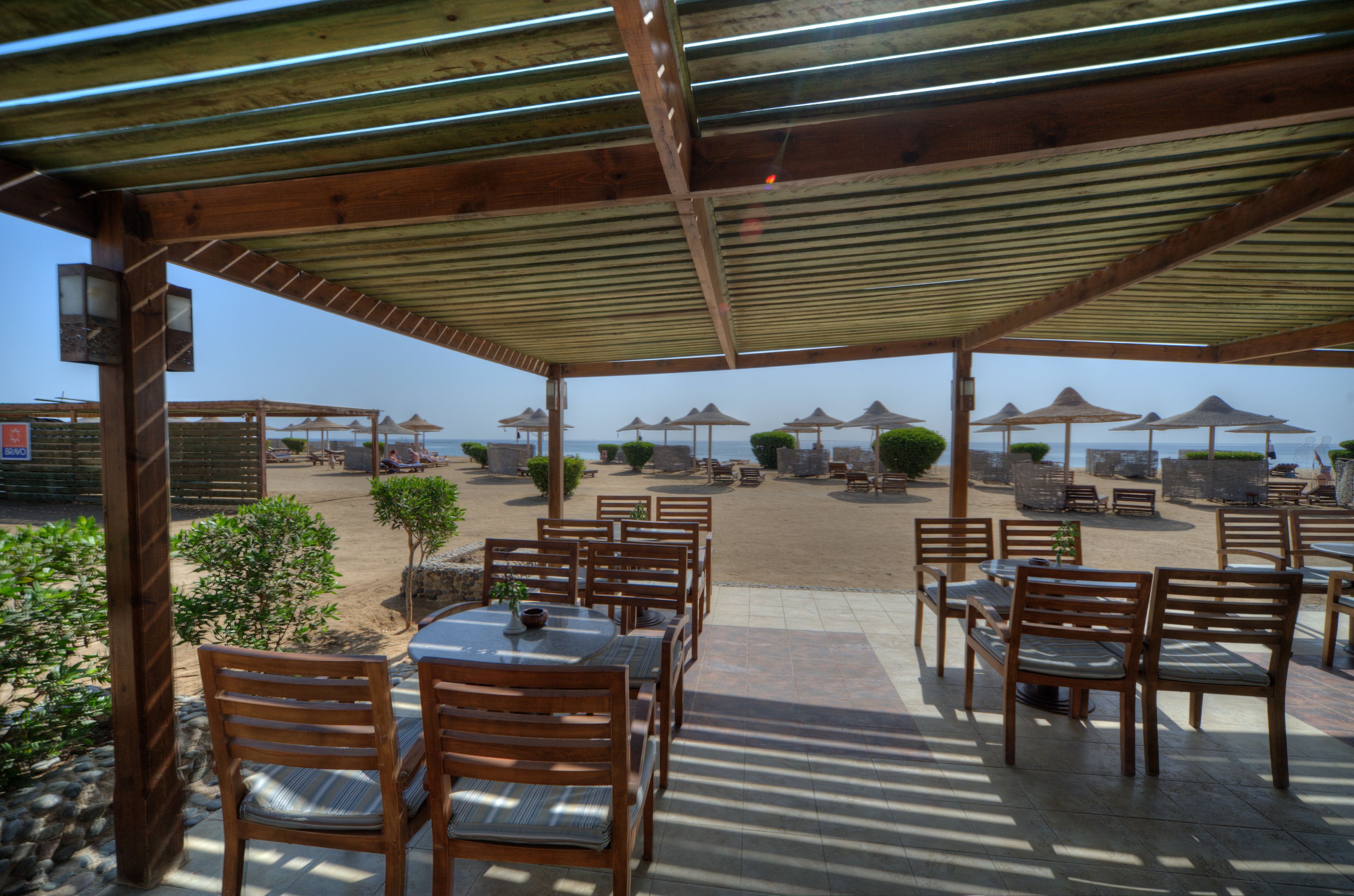 A covered outdoor seating area with wooden tables and chairs, surrounded by umbrellas and small plants.