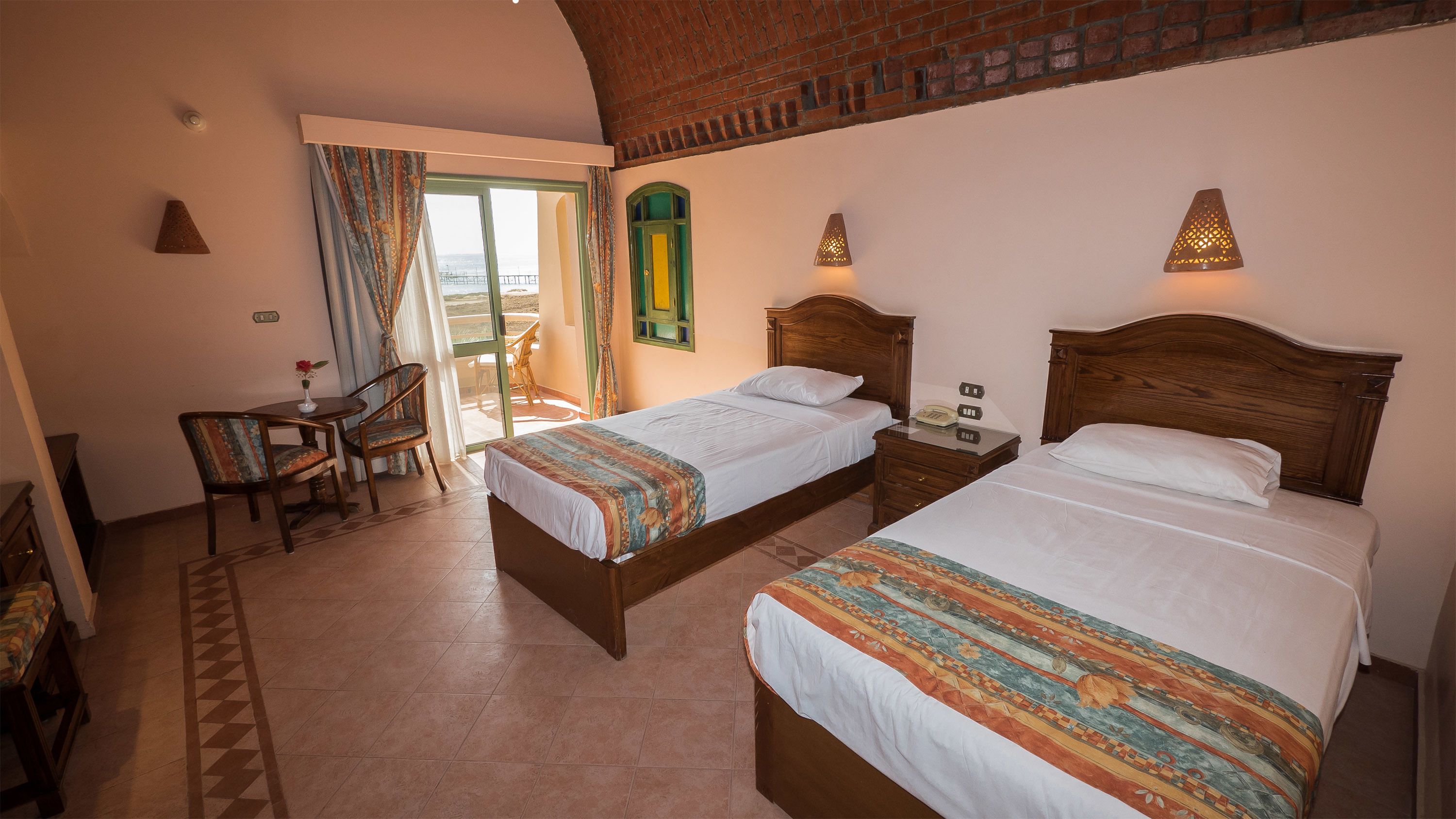 A hotel room with two beds, a balcony, a small table with chairs, and a telephone on the nightstand.