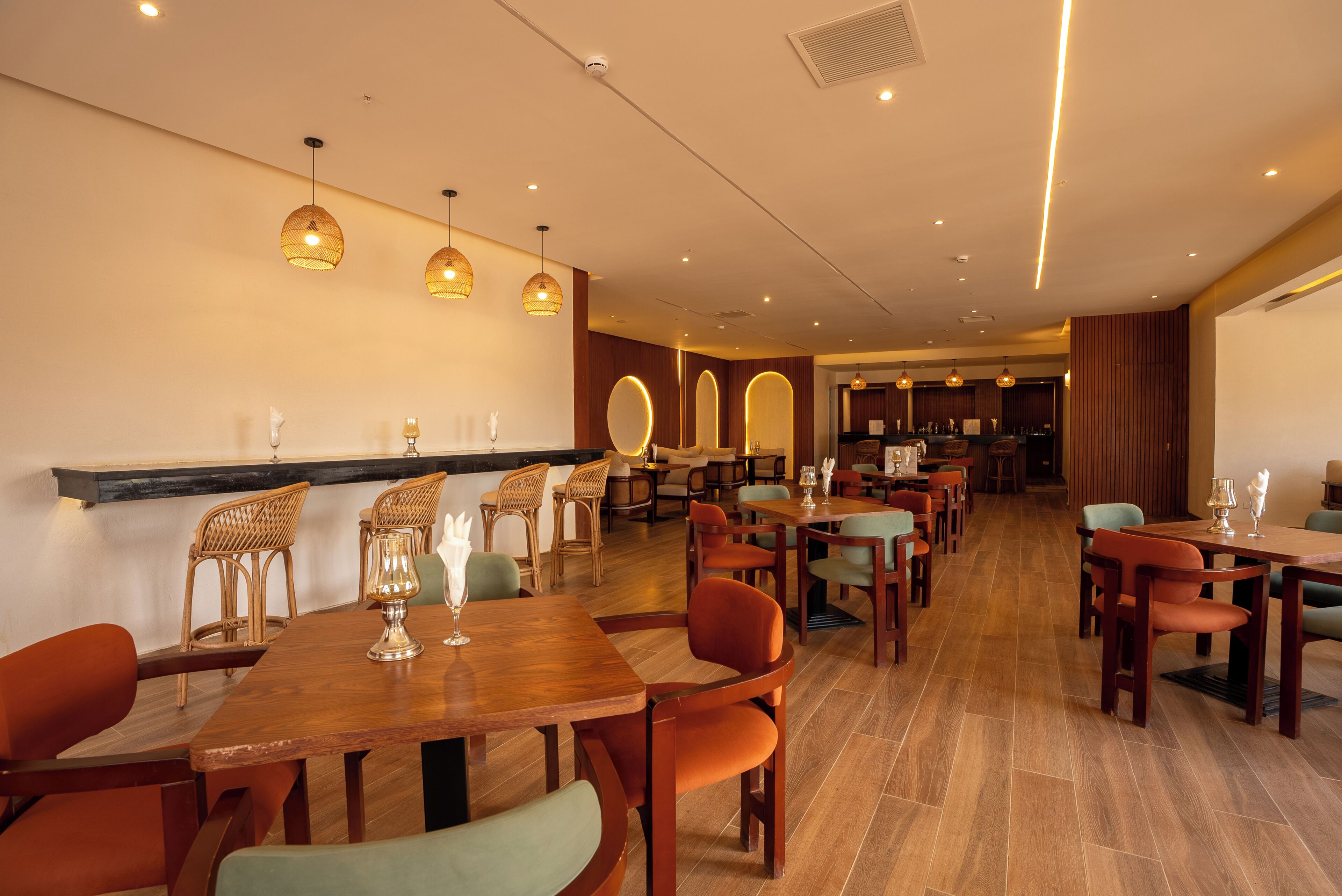 A restaurant with wooden tables and chairs, pendant lighting, and a bar area in the background.