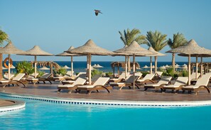 Un espace aménagé au bord de la piscine, avec des chaises longues et des parasols en chaume, offrant une vue sur une mer d’un bleu limpide.