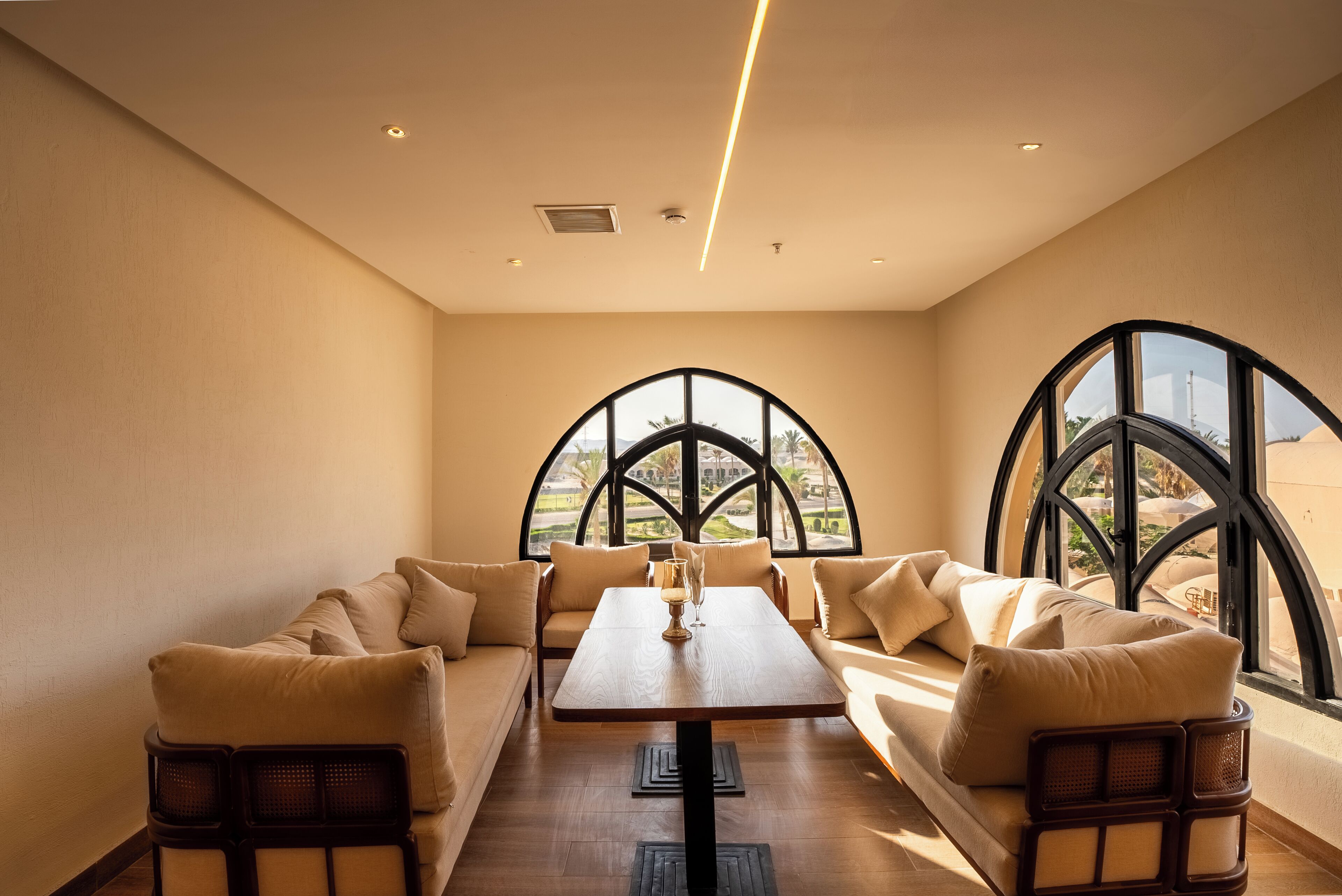 A modern dining area with a long table, cushioned seating, and large arched windows offering a view.