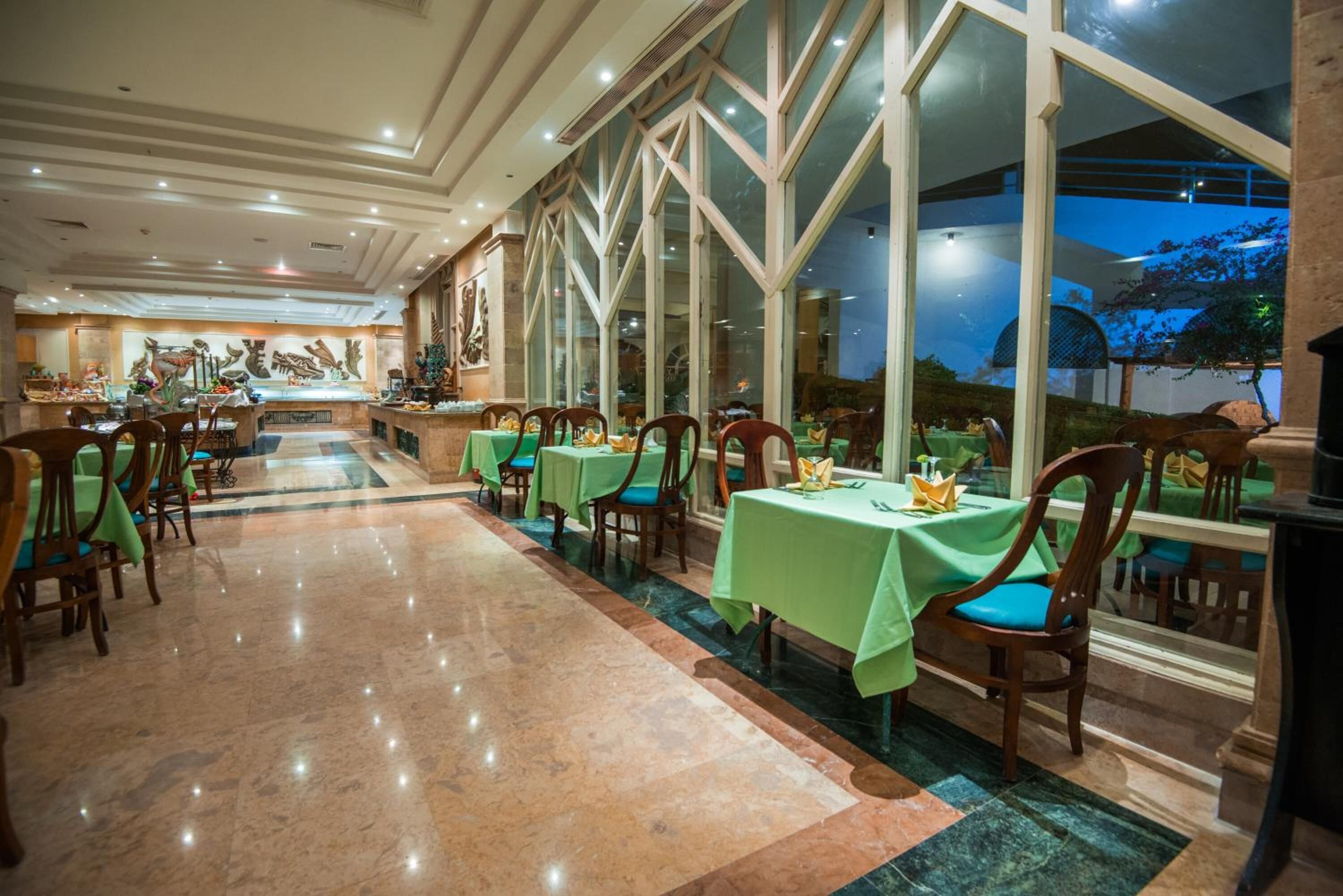 A spacious restaurant with green tablecloths, wooden chairs, and large windows offering a view of the outdoors.