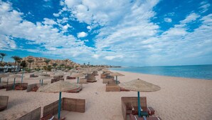 A beach with numerous sun umbrellas and lounge chairs, a clear sky, and a distant view of a coastal town. - Shams Safaga Resort - All inclusive (Safaga)