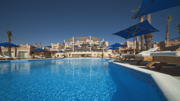 Una piscina con sillones de descanso y sombrillas, rodeada de edificios con balcones y palmeras.
