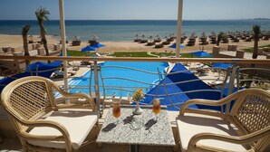 Un balcón con sillas de mimbre y una mesa, con vistas a una piscina y zona de playa con sombrillas y sillones de descanso.