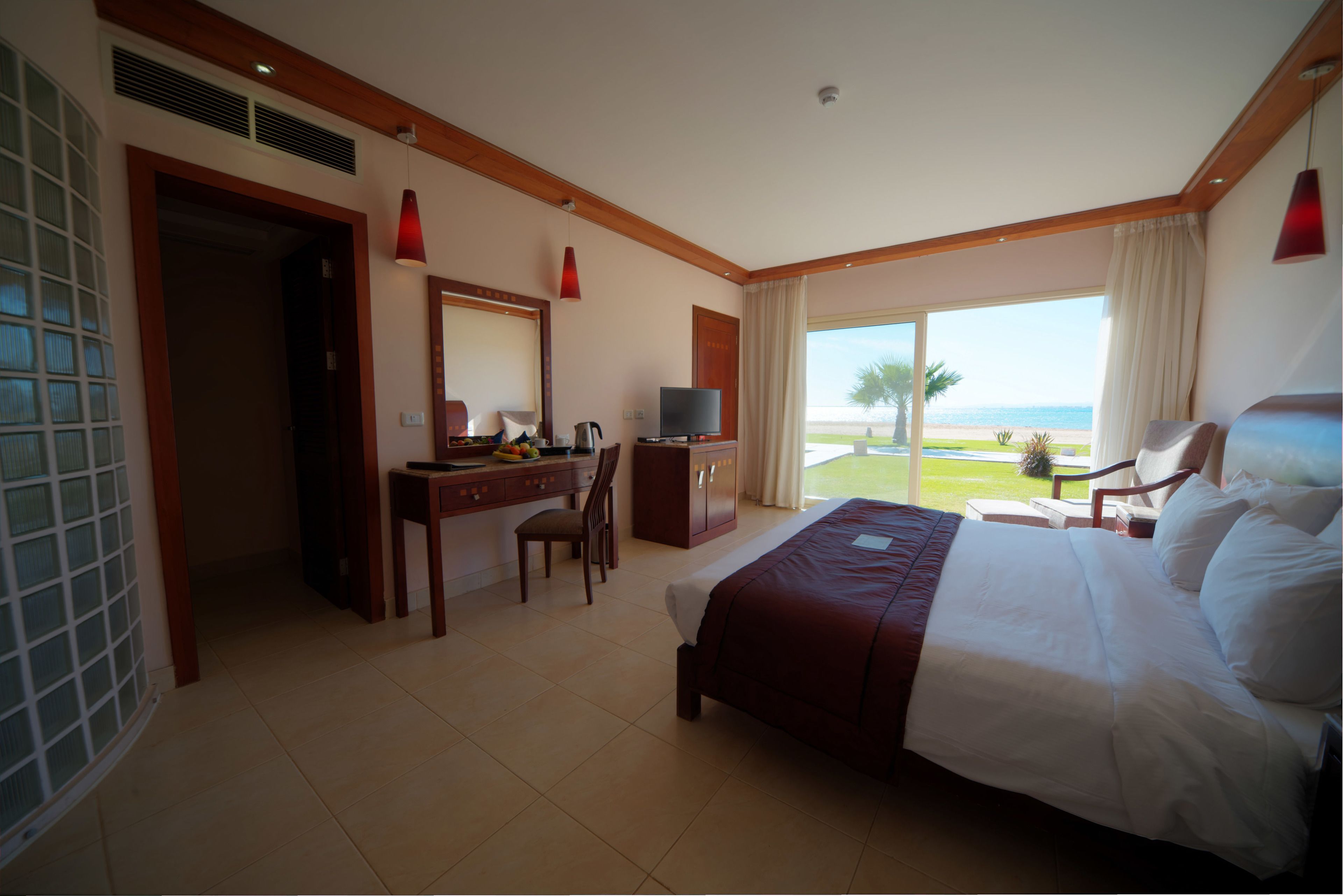 Habitación de hotel con cama, escritorio, silla, televisor y un ventanal con vista a la playa.