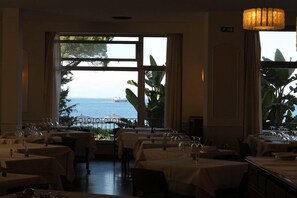 Un comedor con mesas dispuestas para el servicio, amplias ventanas con vista al mar y una lámpara colgante.