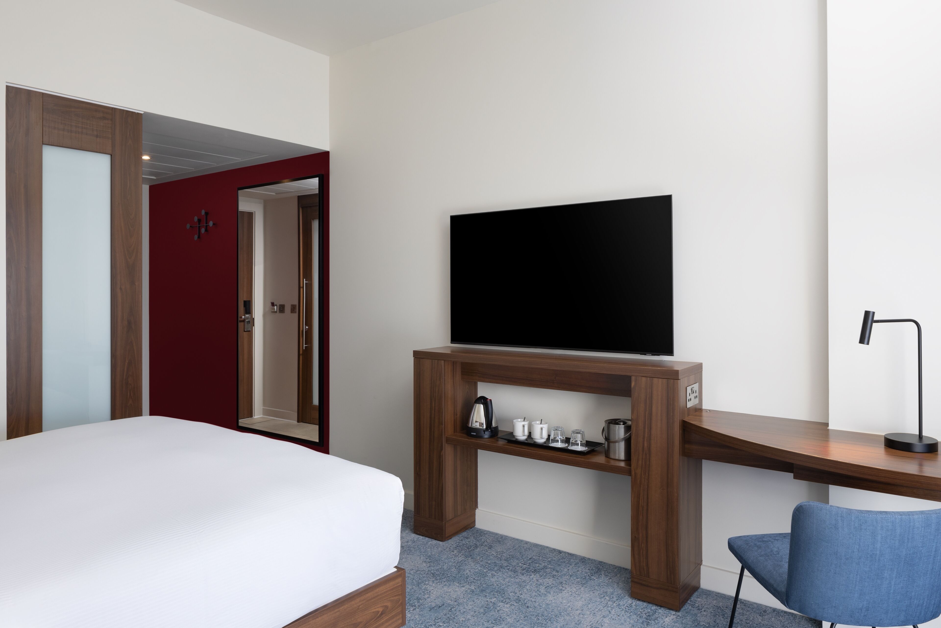 A hotel room with a bed, a TV on a wooden stand, a desk with a chair, and a red door.