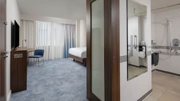 A hotel room with a bed, a desk with a flat-screen TV, a chair, a large mirror, and a shower area.