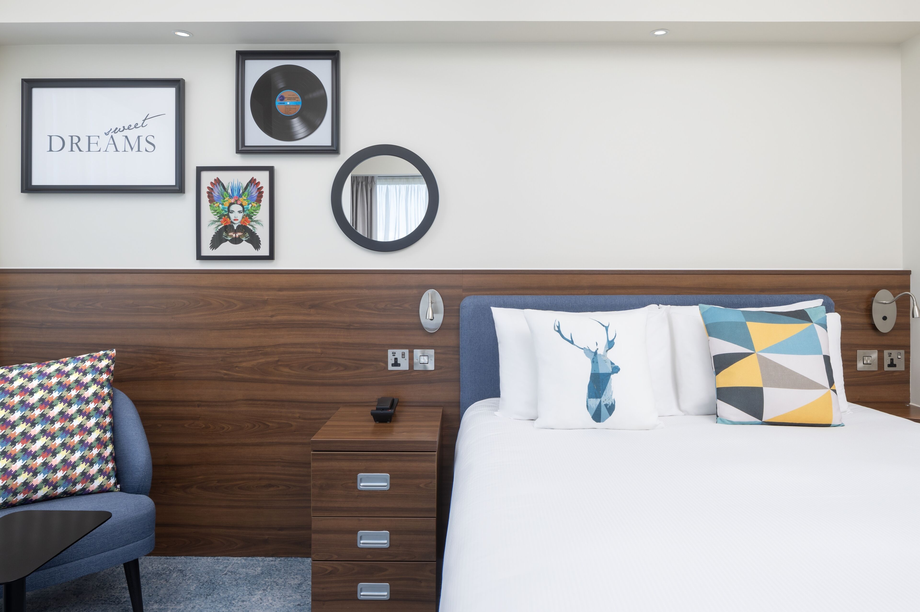 Une chambre d’hôtel avec une tête de lit en bois, un lit agrémenté d’oreillers à motifs, une table de chevet et un mur orné d’œuvres d’art encadrées et d’un miroir.
