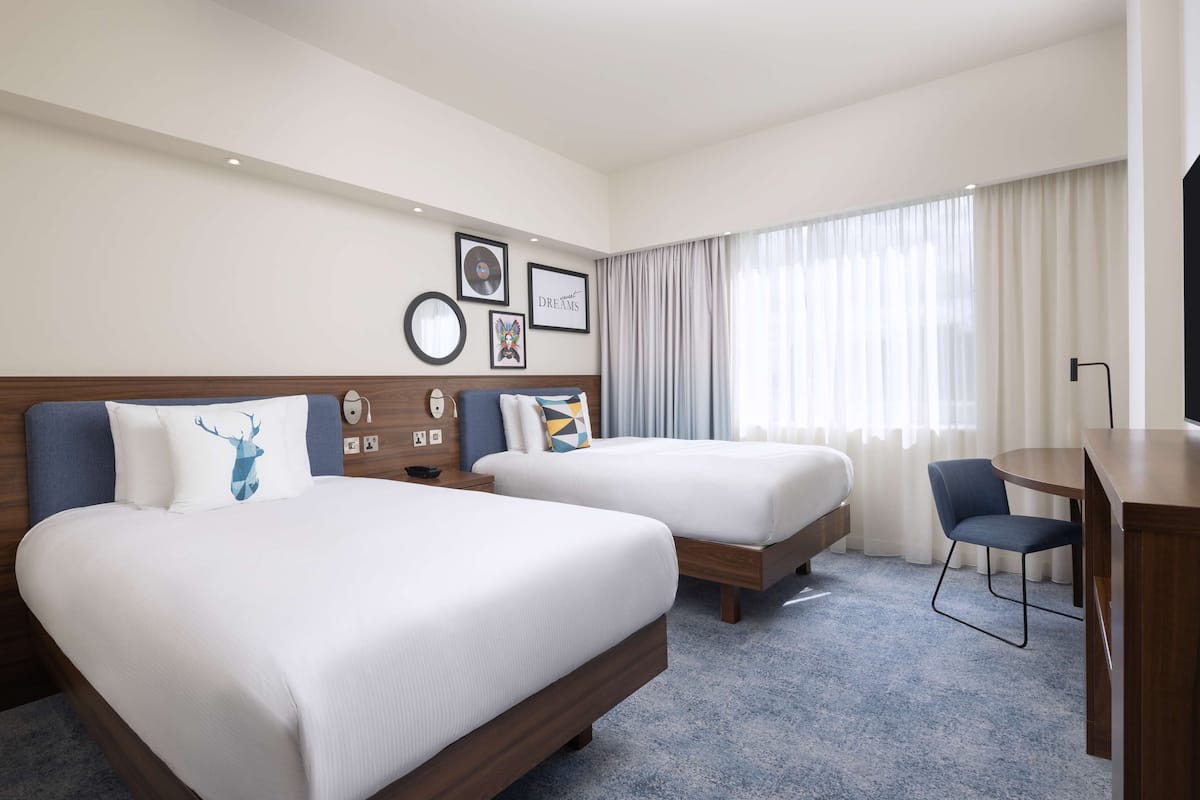 Une chambre d’hôtel avec deux lits, un bureau, une chaise et une grande fenêtre avec des rideaux.