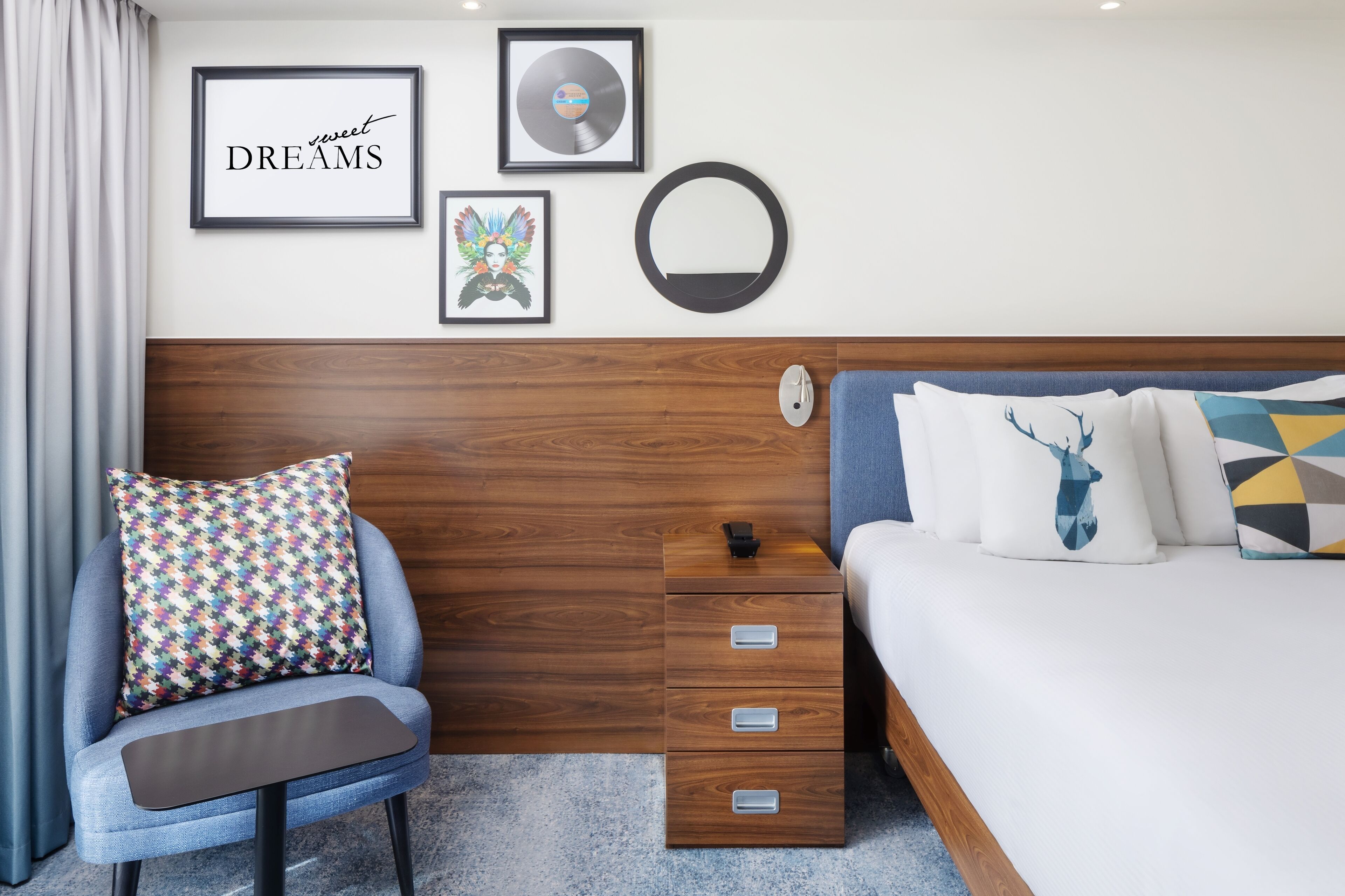 Een moderne slaapkamer met een houten hoofdbord, een bed met gekleurde kussens, een nachtkastje en een stoel met een decoratief kussen.