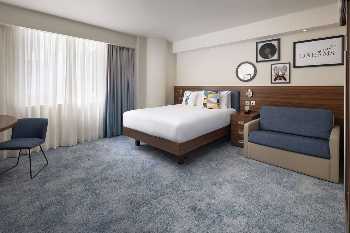 Une chambre d’hôtel avec un lit, un canapé, un bureau et une chaise. On y trouve des rideaux, une horloge murale et des tableaux encadrés accrochés au mur.