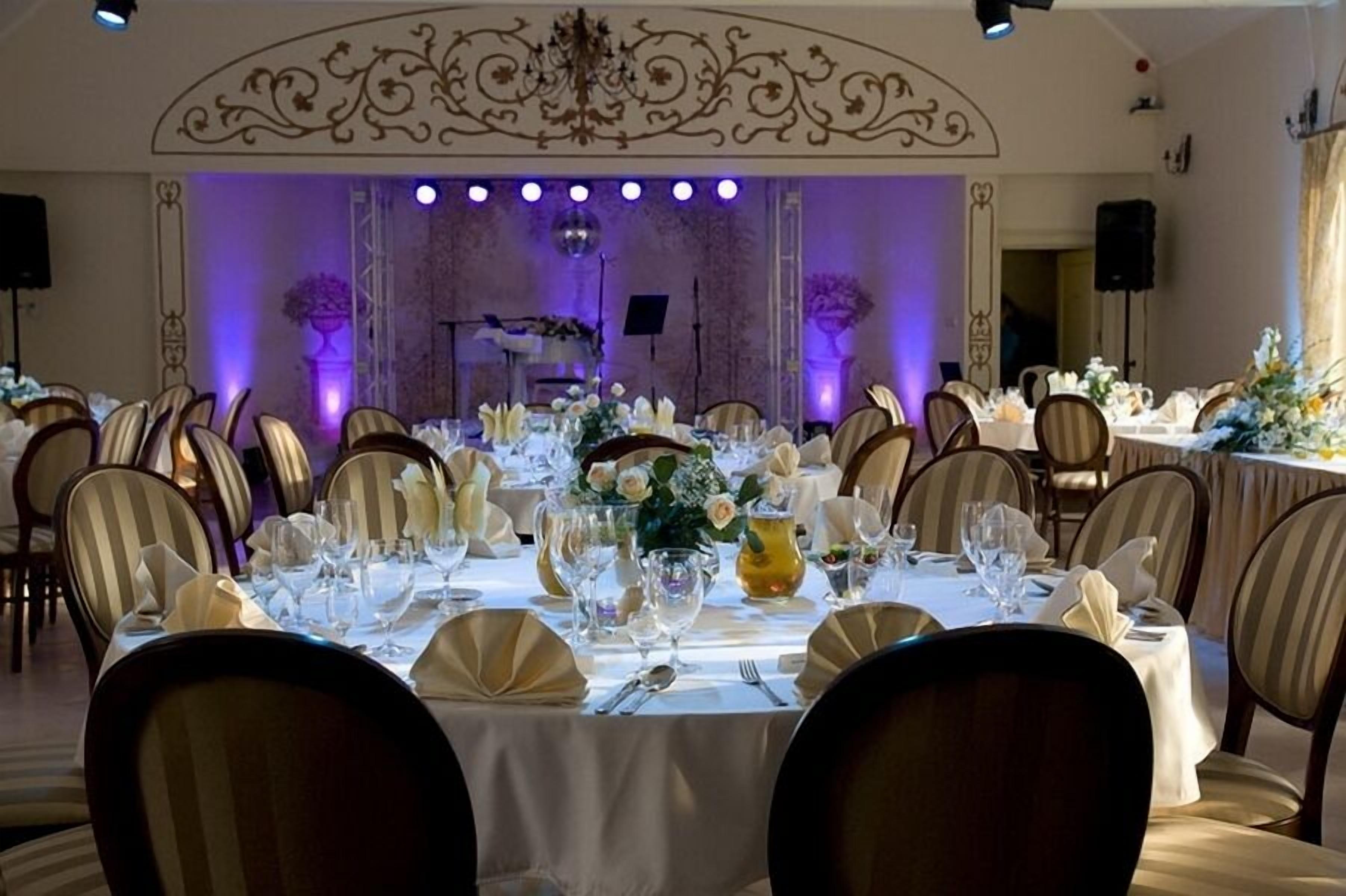 A banquet hall with round tables set for a formal event, featuring white tablecloths, floral centerpieces, and striped chairs.