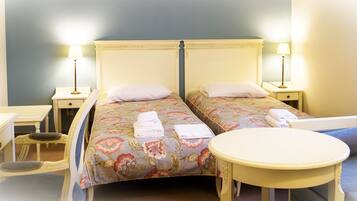 Habitación de hotel con dos camas individuales, una mesita redonda pequeña, un escritorio con una silla y una mesita de noche con lámpara.