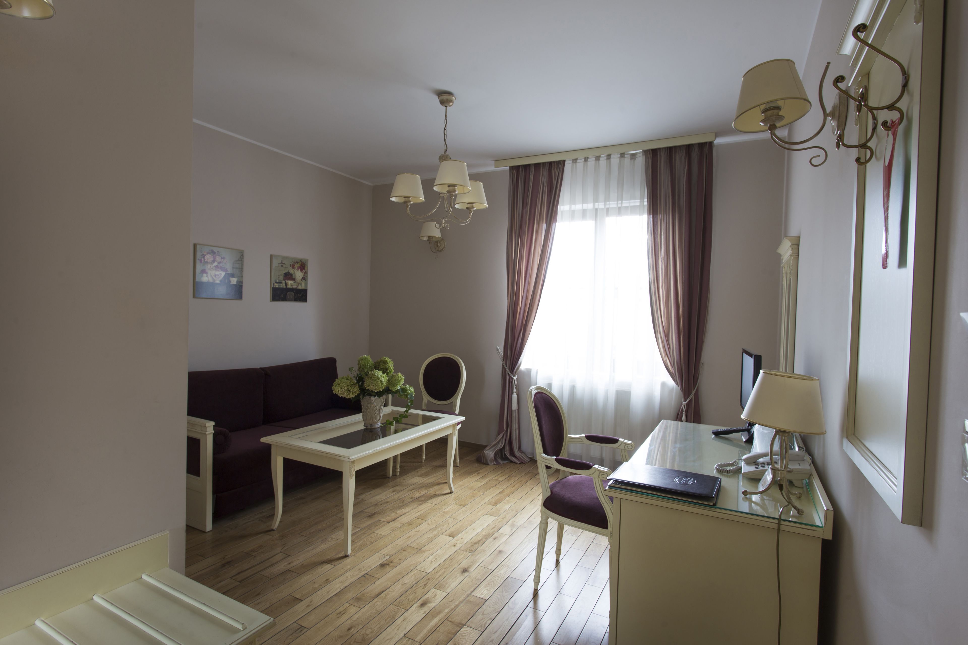 A hotel room with a wooden floor, a white sofa, a dining table with chairs, a desk with a lamp, and a large window with curtains.