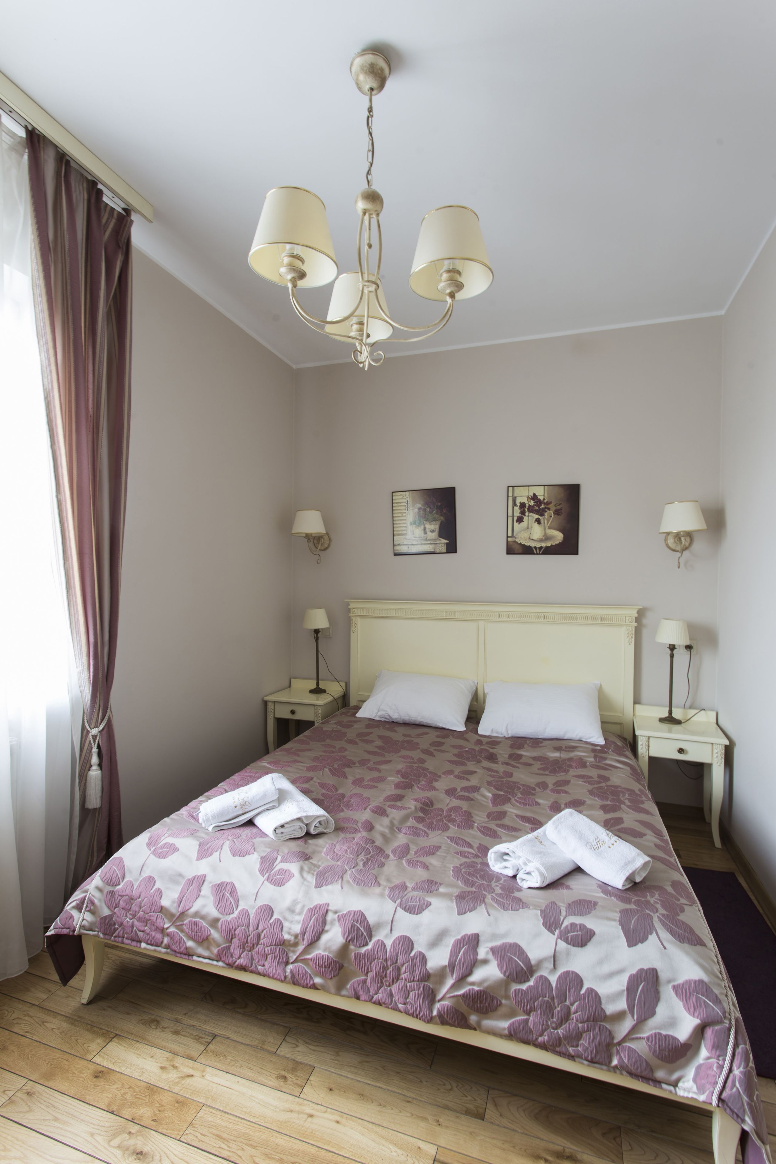 Una cama bien tendida con ropa de cama floral, un candelabro y lámparas de noche en una habitación con suelo de madera y una ventana con cortinas.