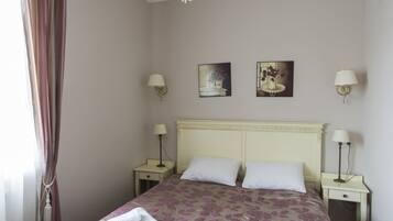 Una cama bien tendida con ropa de cama floral, un candelabro y lámparas de noche en una habitación con suelo de madera y una ventana con cortinas.