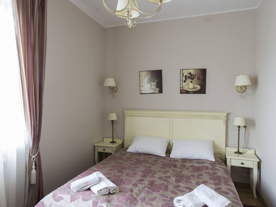 Una cama bien tendida con ropa de cama floral, un candelabro y lámparas de noche en una habitación con suelo de madera y una ventana con cortinas.