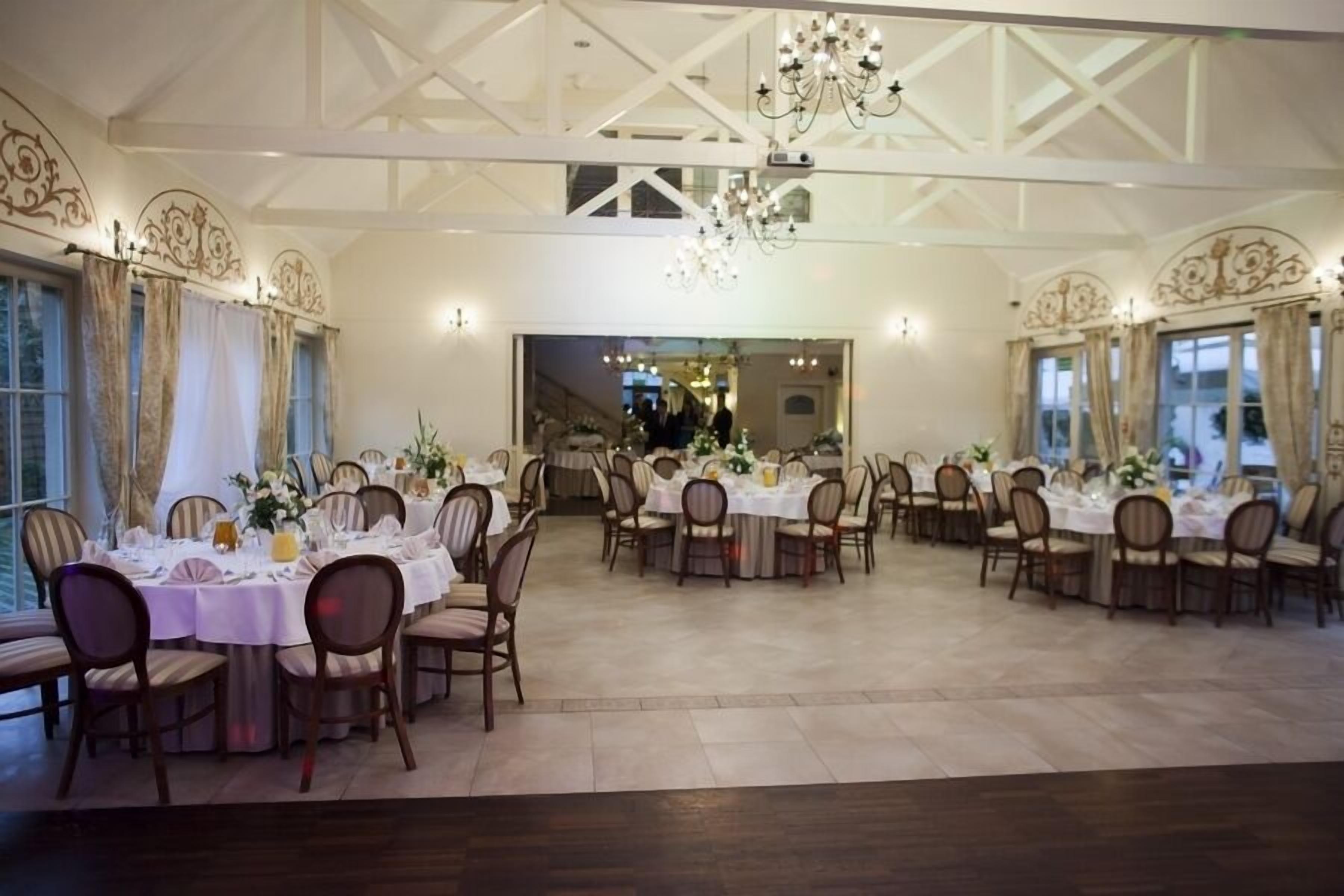 Une grande salle à manger avec de nombreuses tables rondes dressées pour le repas, dotée d’un plafond haut avec des poutres apparentes et des lustres.