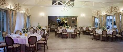 Une grande salle à manger avec de nombreuses tables rondes dressées pour le repas, dotée d’un plafond haut avec des poutres apparentes et des lustres.
