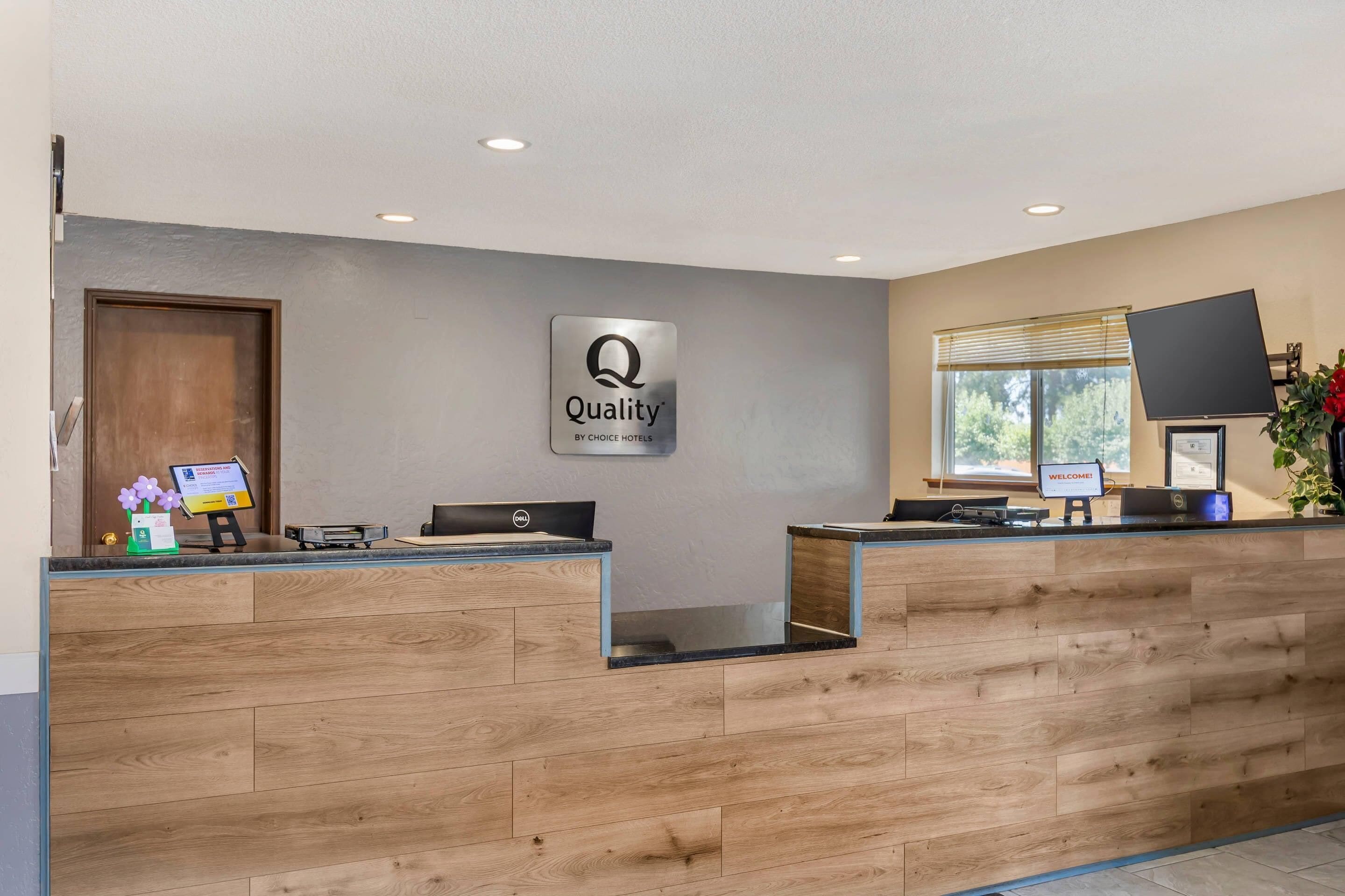 Een receptiebalie van een hotel met een bord waarop staat 'Quality by Choice Hotels', een computer en een raam met jaloezieën.