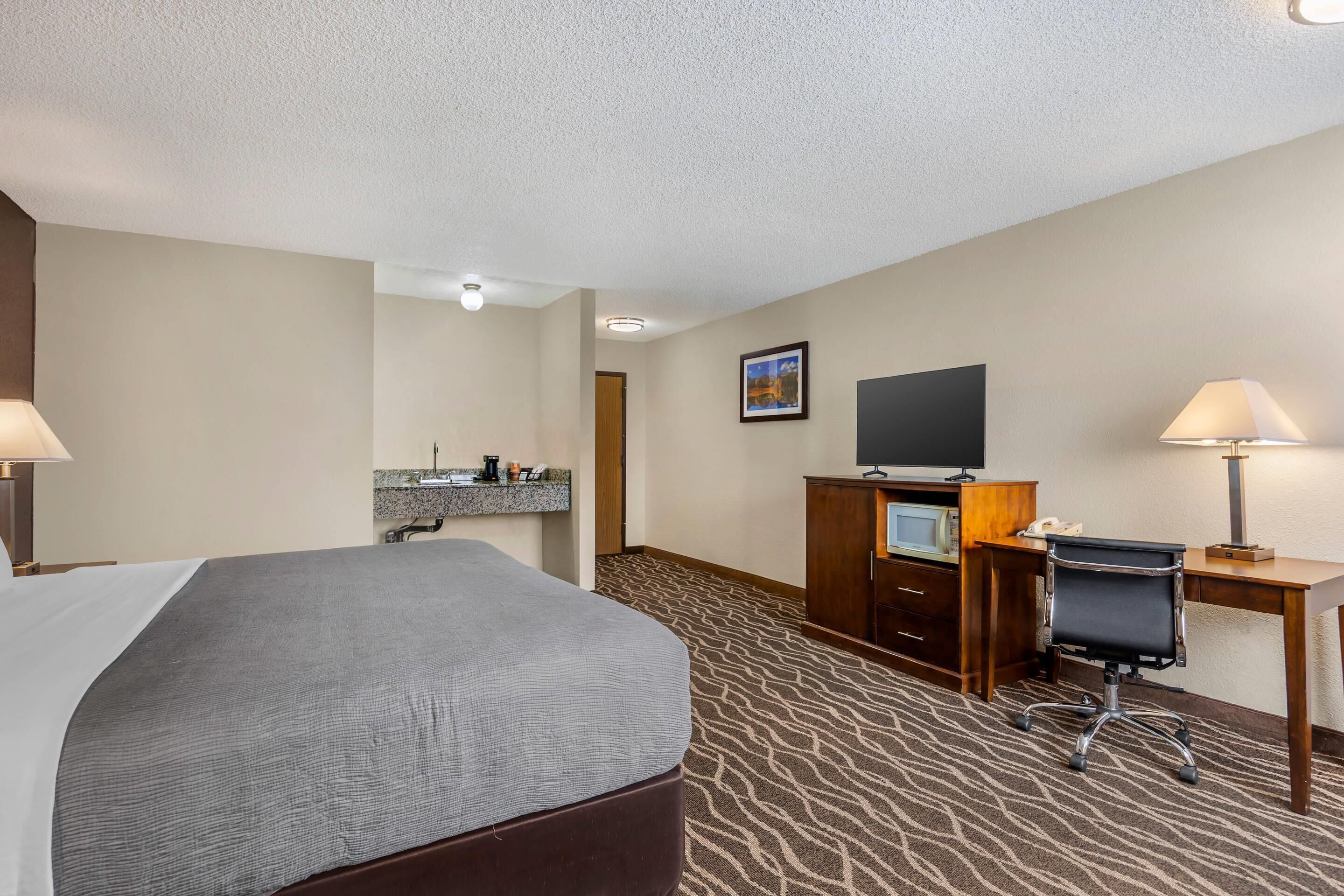 A hotel room with a bed, desk, chair, TV, and a small kitchenette area.