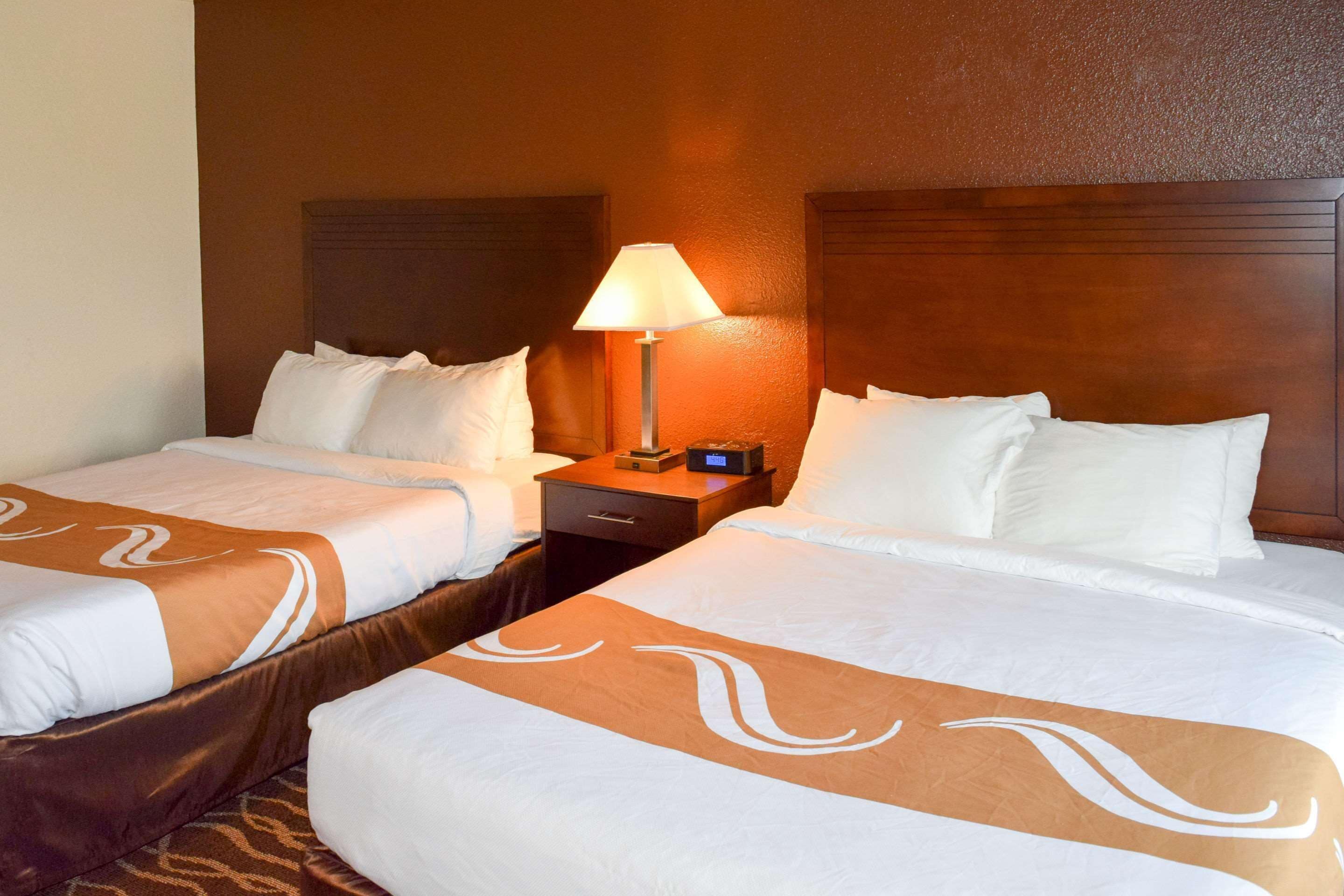 Ein Hotelzimmer mit zwei Betten, beide mit weißen Laken und braunen Zierleisten. Dazwischen steht ein Nachttisch mit einer Lampe.