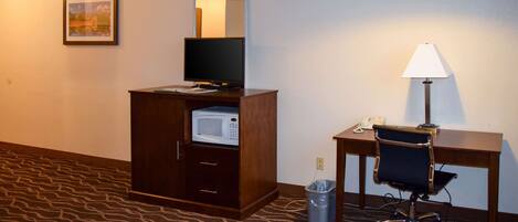 A hotel room with a desk, chair, microwave, trash can, and a lamp.