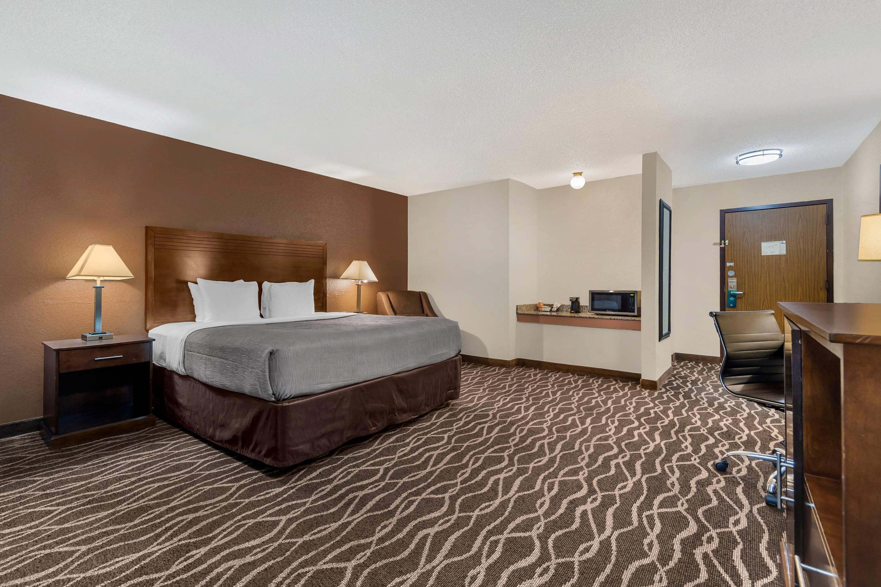A hotel room with a bed, two bedside lamps, a desk with a chair, a microwave, and a door with a sign.