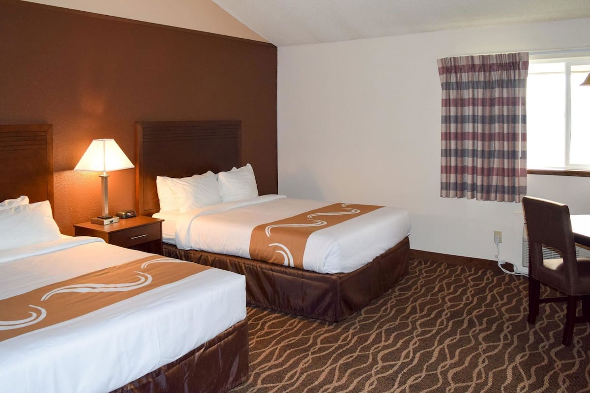 A hotel room with two beds, a desk, a chair, a lamp, and a window with curtains.