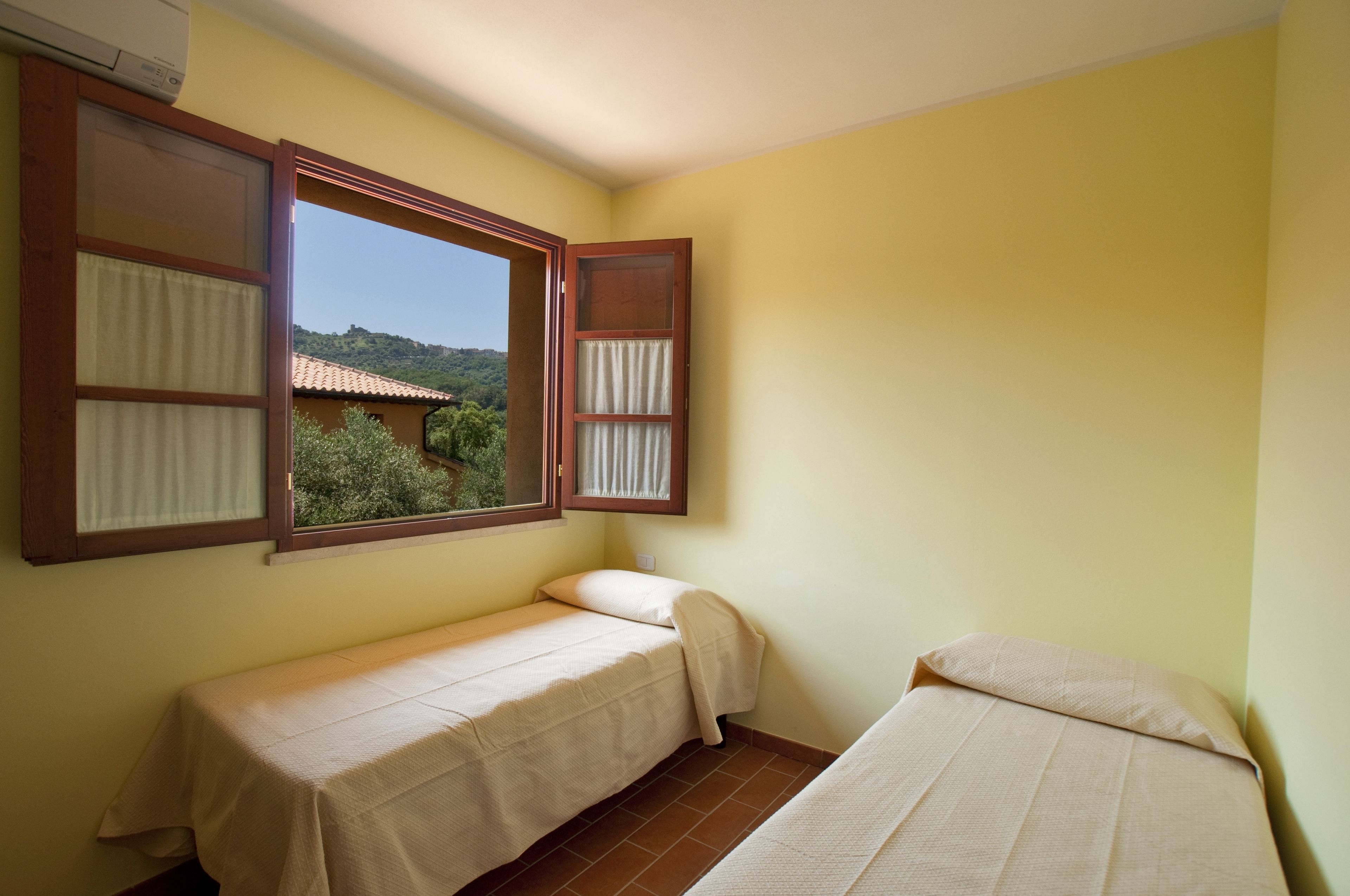 A small room with two beds, a window with curtains, and a view of greenery outside.
