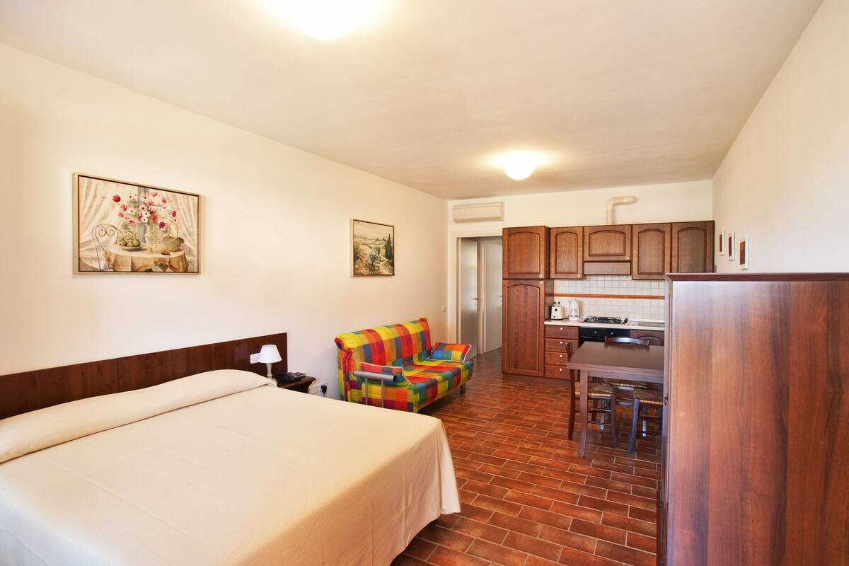 Ein Hotelzimmer mit Bett, einem bunten Sessel, einem Esstisch und einer Küchenzeile.
