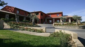 A two-story house with a red facade, a stone pathway leading to the entrance, and a landscaped garden with various plants and flowers. - Borgo Etrusco S.p.A. (Scarlino)