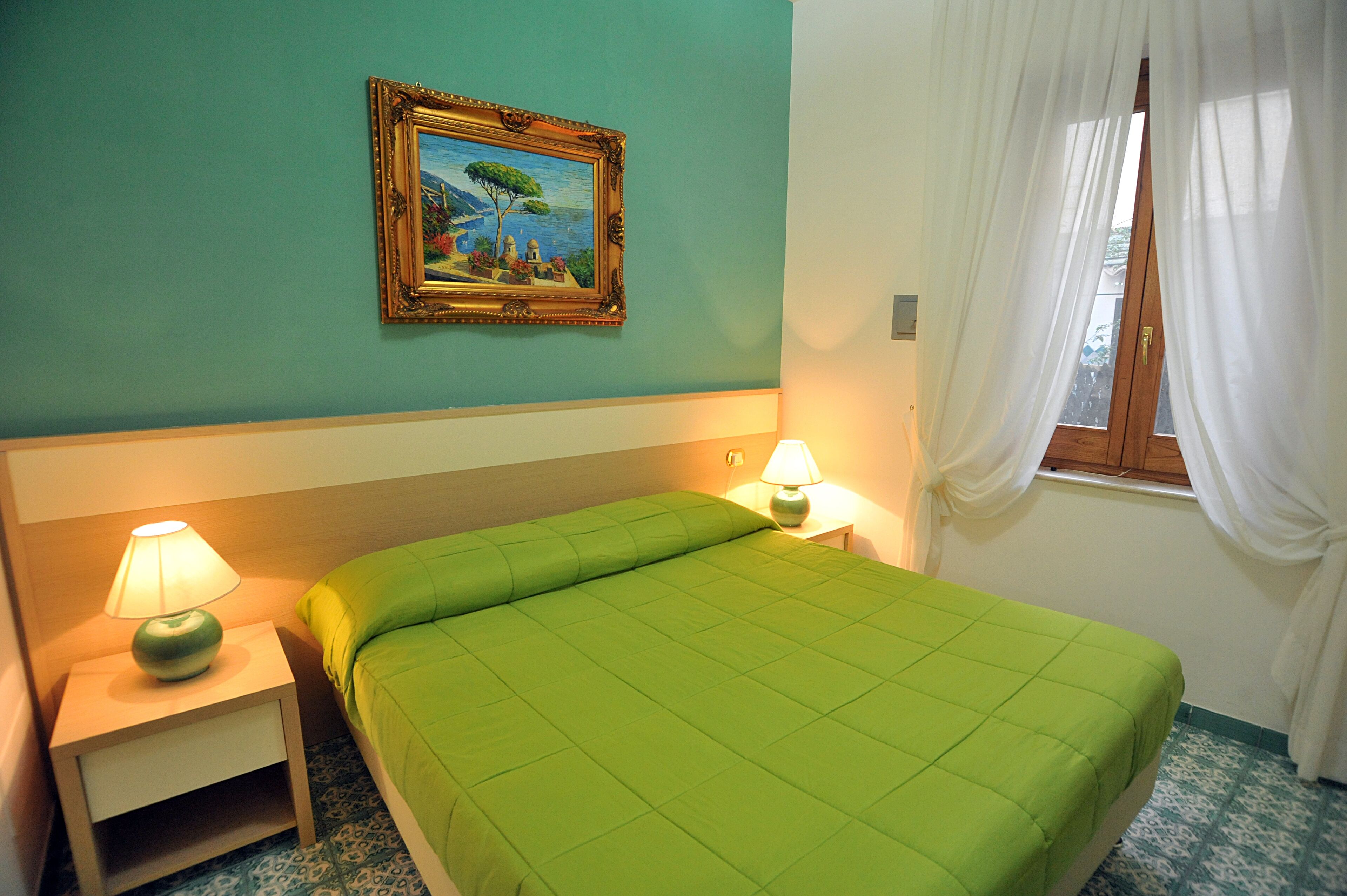 A room with a bed, two bedside lamps, a framed painting on the wall, and a window with curtains.