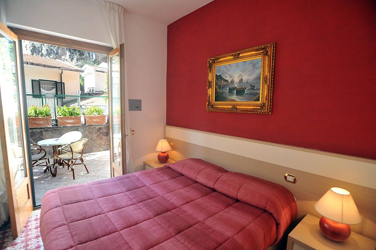 Ein Hotelzimmer mit Bett, einem Gemälde an der Wand, einem Tisch mit Stühlen und Blick auf den Balkon.